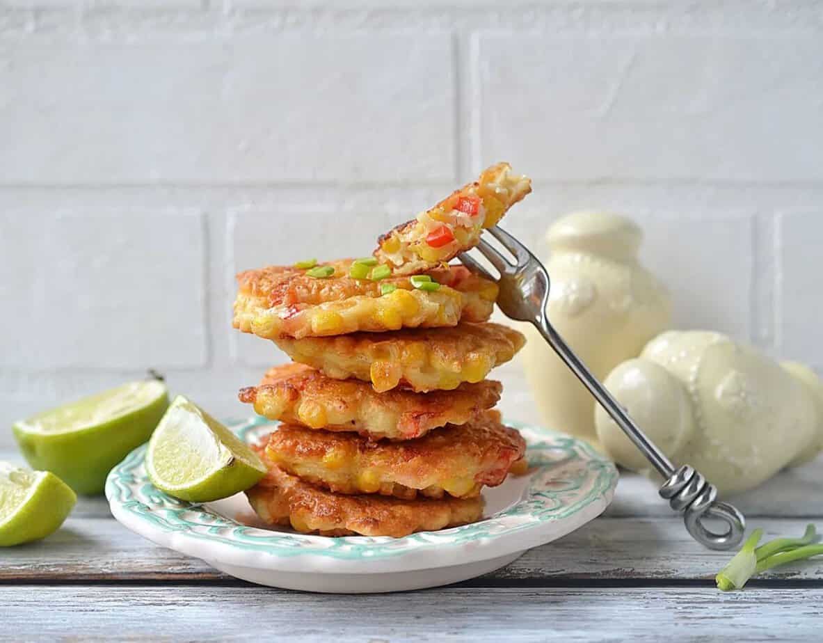 A stack of corn fritters on a plate with lime wedges, garnished with chopped green onions; a fork holds one fritter—perfect for anyone who loves cheesy recipes.