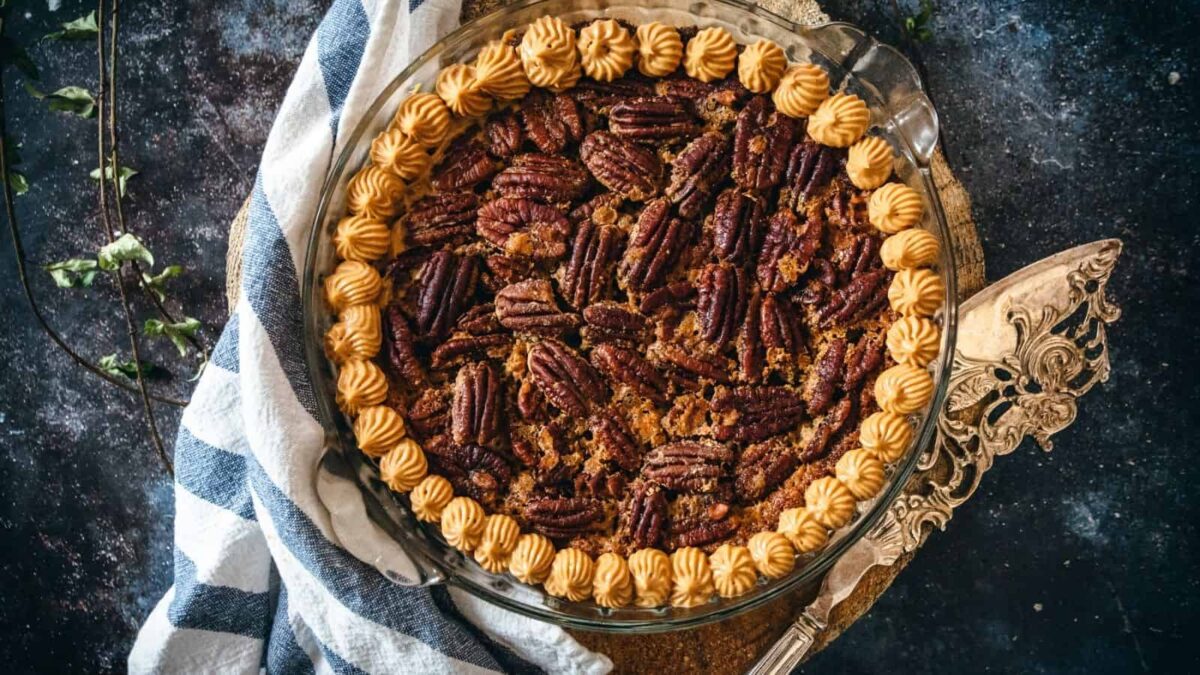 A pecan pie in a glass dish, topped with whole pecans and a piped border of cream, sits on a decorative tray next to a striped cloth—perfect for your holiday lineup of delicious pecan recipes.