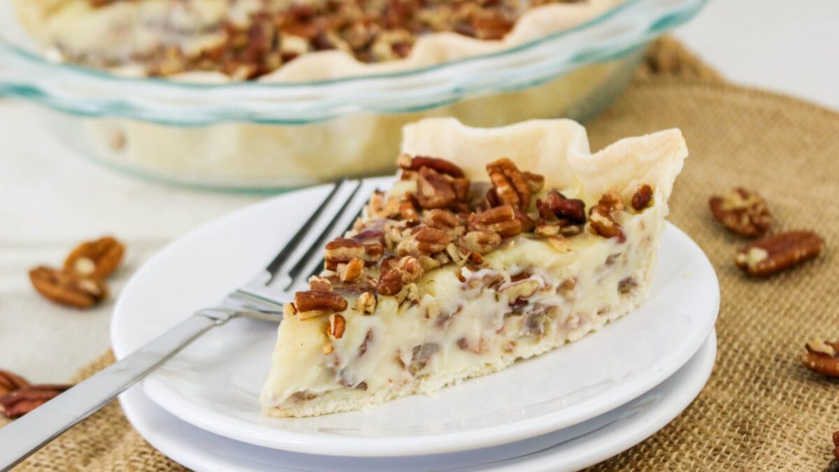 A slice of creamy pecan pie on a white plate with a fork, set on a burlap surface—perfect for your holiday recipes lineup; the whole pie sits in a glass dish in the background.