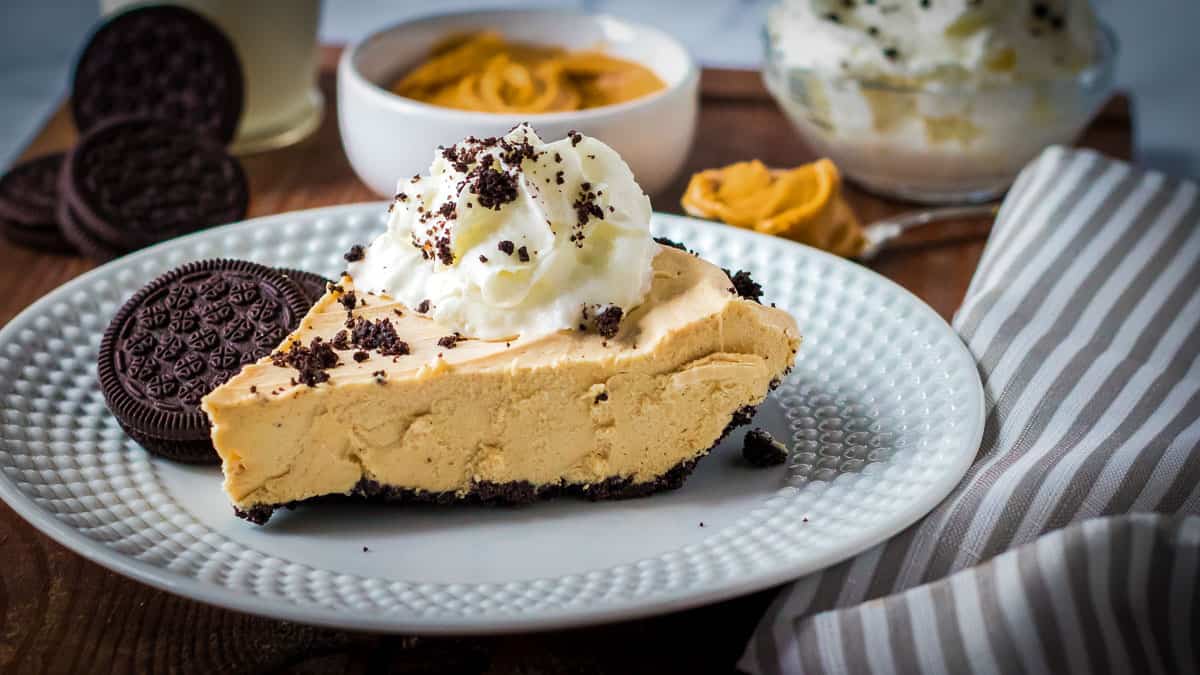 A slice of peanut butter pie with a cookie crust, topped with whipped cream and cookie crumbs, served on a white plate with two chocolate sandwich cookies—a must-try for fans of delicious desserts.
