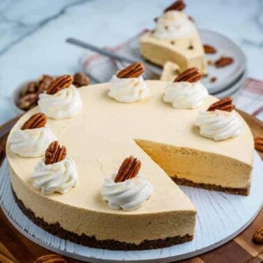A delicious pumpkin cheesecake with a slice removed, topped with whipped cream and pecans, sits on a wooden serving board against a marble surface—perfect for lovers of delicious desserts.