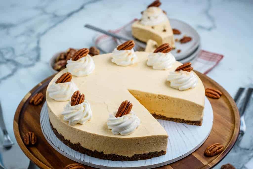 A delicious pumpkin cheesecake with a slice removed, topped with whipped cream and pecans, sits on a wooden serving board against a marble surface—perfect for lovers of delicious desserts.