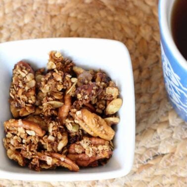 A white bowl filled with granola and nuts sits on a woven placemat next to a blue-patterned cup of dark liquid—an inviting setup for easy vegan breakfast lovers.