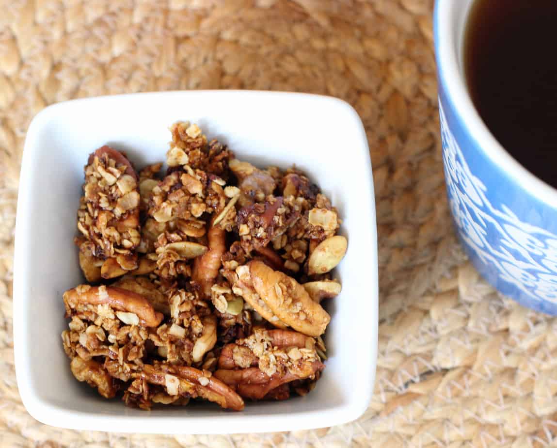 A white bowl filled with granola and nuts sits on a woven placemat next to a blue-patterned cup of dark liquid—an inviting setup for easy vegan breakfast lovers.