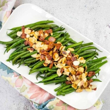 A rectangular white platter with cooked green beans topped with crispy bacon pieces and sliced almonds, placed on a floral-patterned cloth—perfect for Thanksgiving recipes.