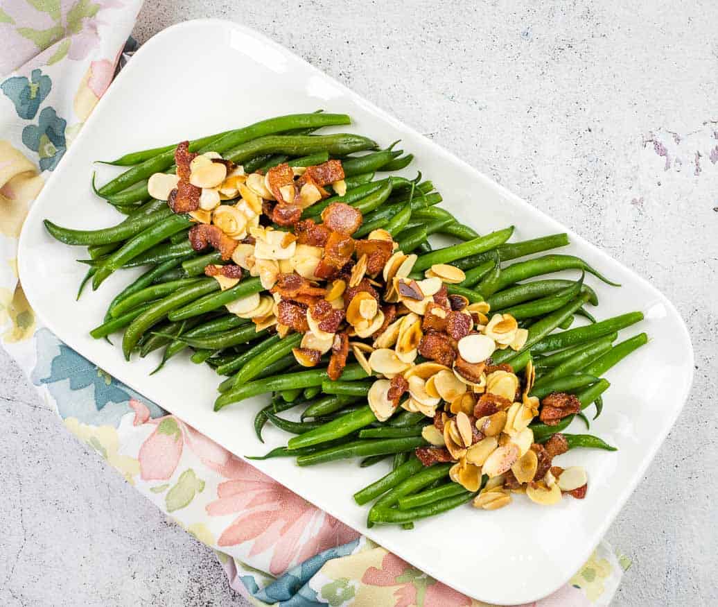 A rectangular white platter with cooked green beans topped with crispy bacon pieces and sliced almonds, placed on a floral-patterned cloth—perfect for Thanksgiving recipes.