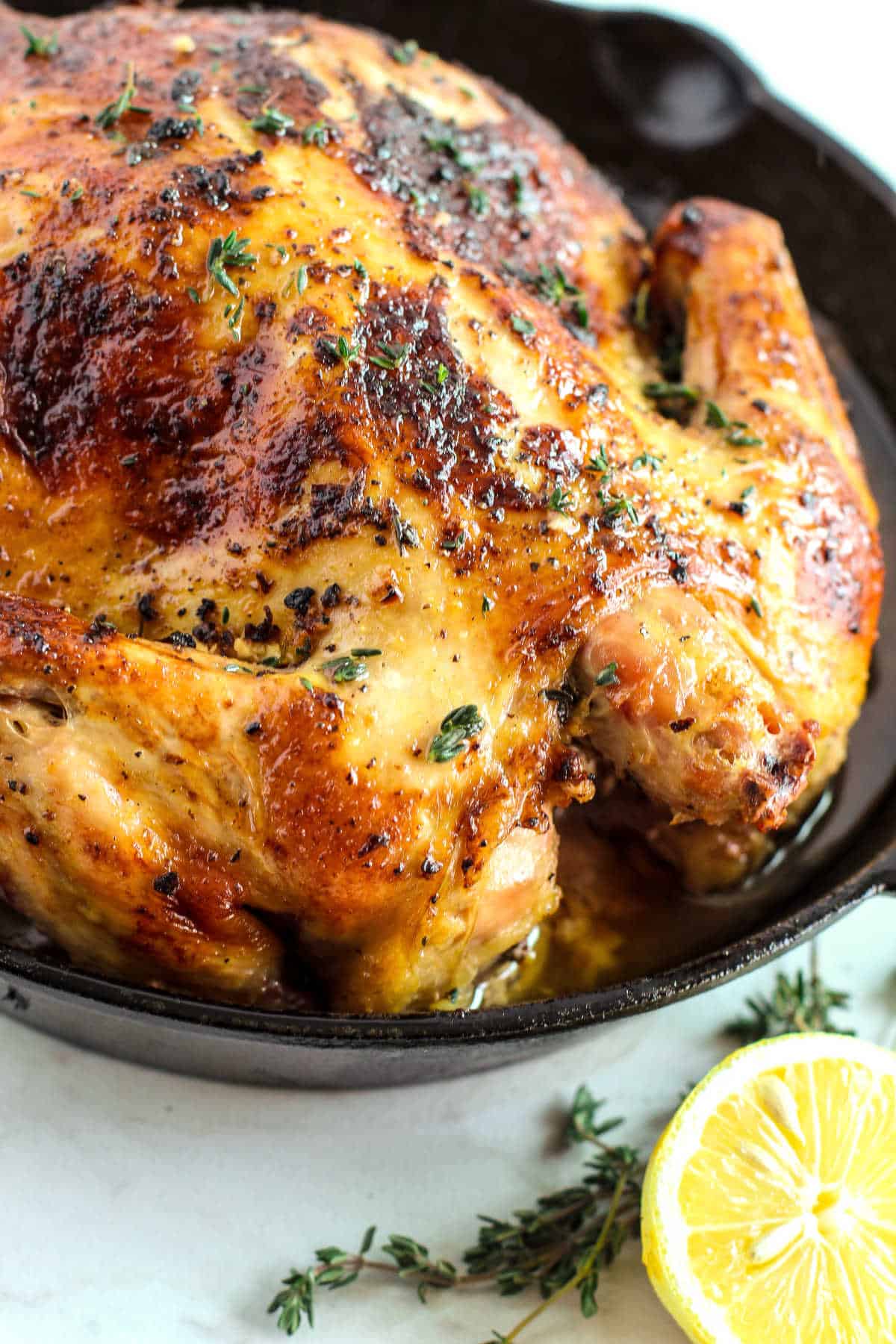 A whole roasted chicken with golden, crispy skin sits in a cast iron pan, garnished with fresh herbs and next to a sliced lemon—perfect for Thanksgiving recipes or any special gathering.