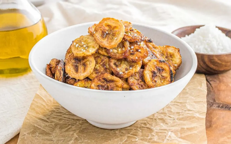 A white bowl filled with golden-brown fried plantain chips, perfect for vegan recipes, with a bottle of oil and a small bowl of salt in the background.