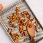 A baking sheet with roasted sweet potato cubes spread out, some being scooped with a wooden spoon, on a light surface.