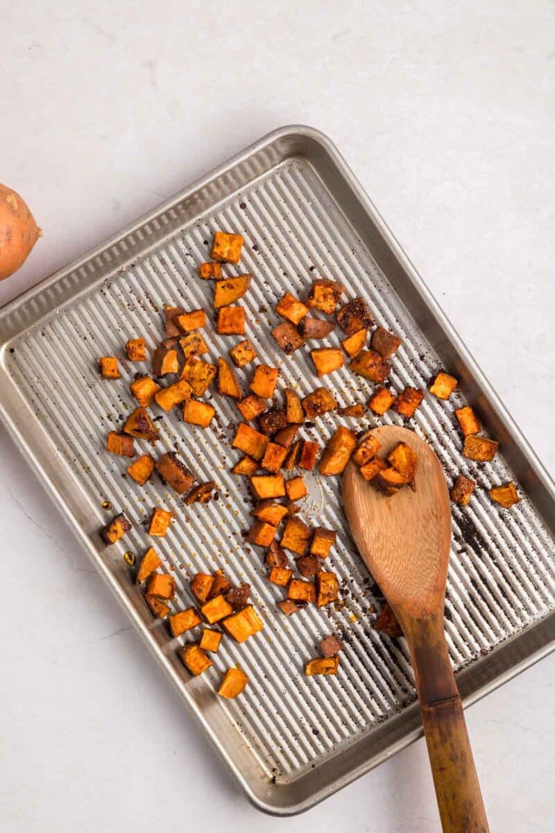 A baking sheet with roasted sweet potato cubes spread out, some being scooped with a wooden spoon, on a light surface.