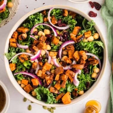 A bowl of salad with kale, roasted sweet potatoes, chickpeas, red onion, dried cranberries, and pumpkin seeds, next to a green napkin, salad dressing, fork, and wooden salad tongs.
