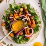 A hand drizzles dressing over a salad with kale, roasted sweet potatoes, chickpeas, red onion, and pumpkin seeds on a patterned plate.