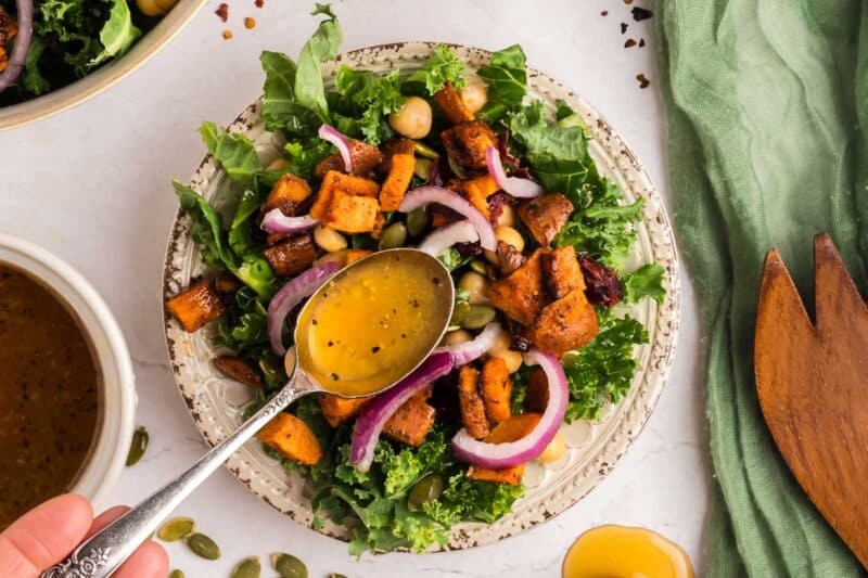 A hand drizzles dressing over a salad with kale, roasted sweet potatoes, chickpeas, red onion, and pumpkin seeds on a patterned plate.
