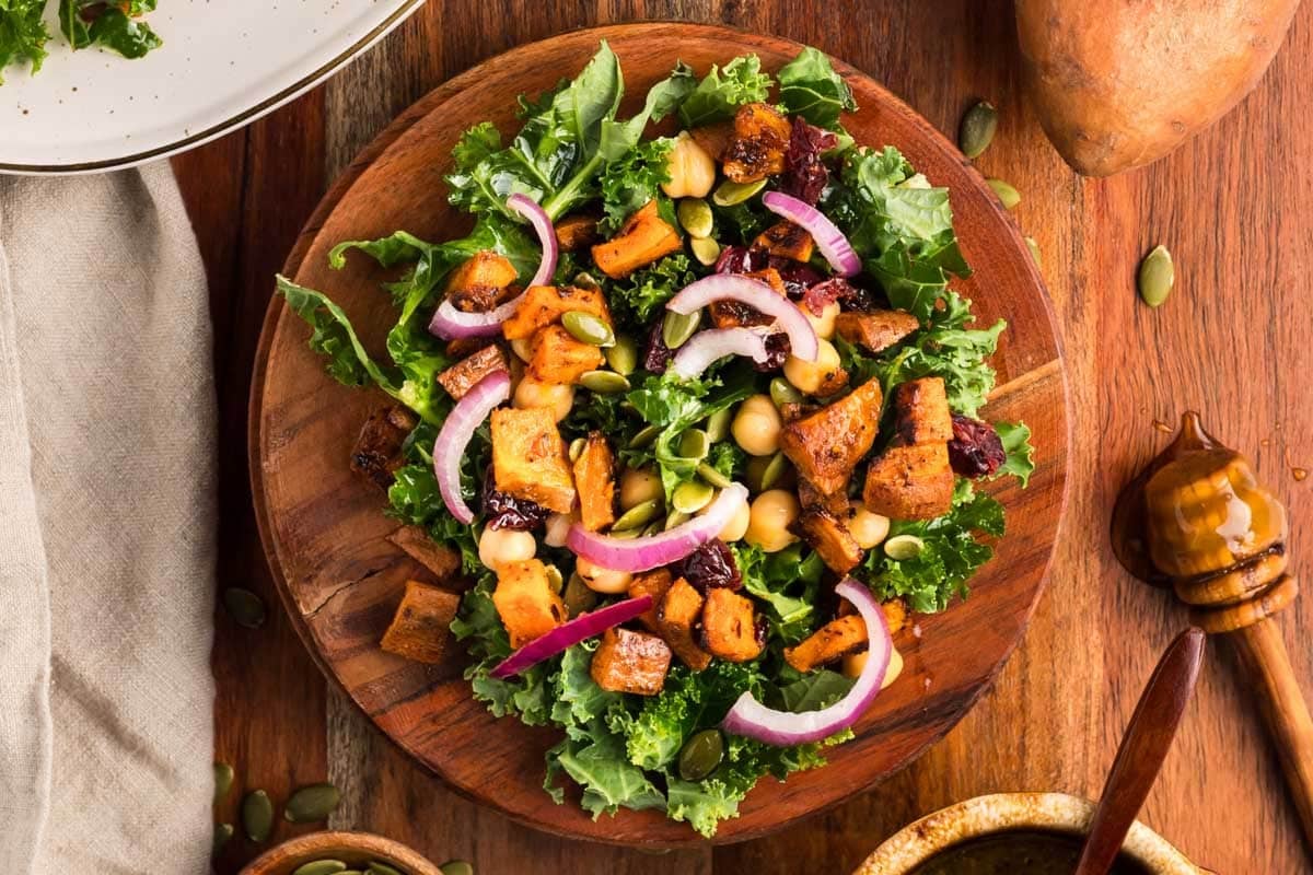 A salad with kale, roasted sweet potatoes, chickpeas, red onion slices, dried cranberries, and pumpkin seeds on a wooden plate.