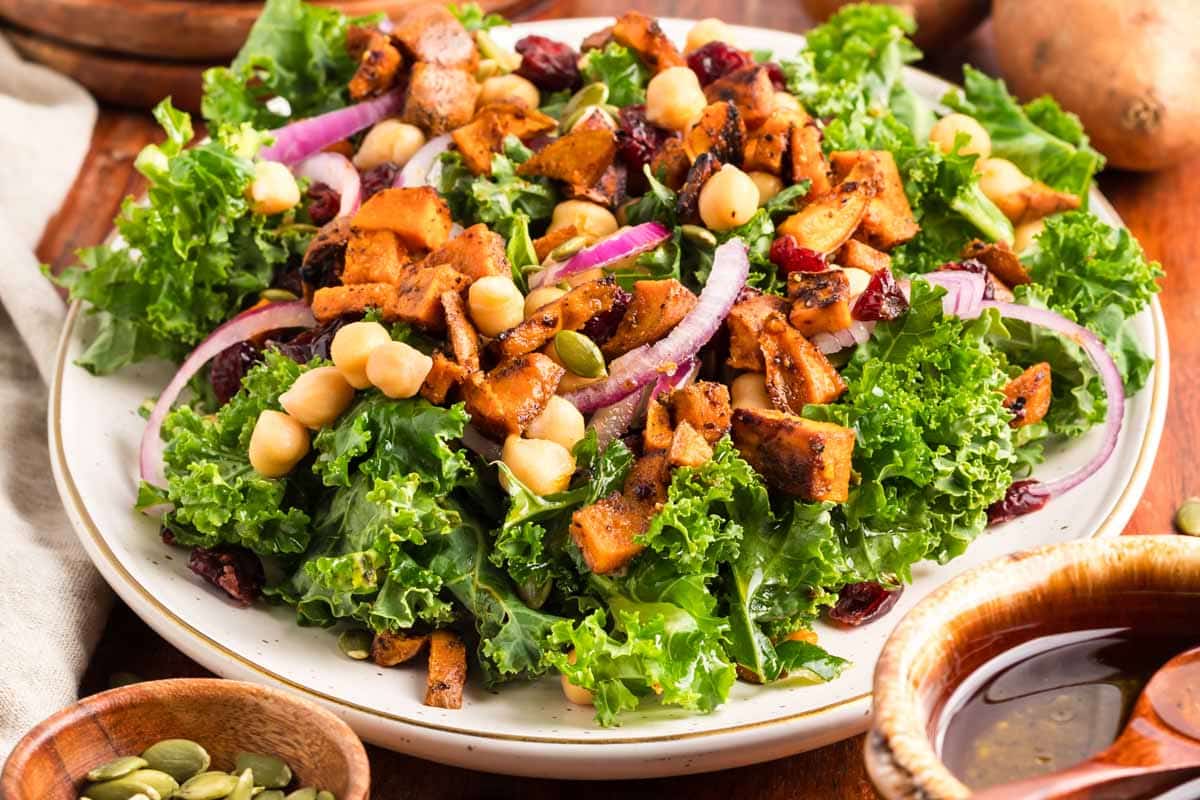 A plate of kale salad topped with roasted sweet potatoes, red onion slices, chickpeas, dried cranberries, and pumpkin seeds.