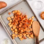 Cubed sweet potatoes seasoned with spices are spread on a baking sheet, surrounded by empty glass bowls, a wooden spoon, and a sweet potato on a white surface.