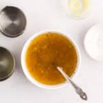 A bowl of vinaigrette with a spoon inside, surrounded by empty measuring cups and a small dish of mustard on a white surface.
