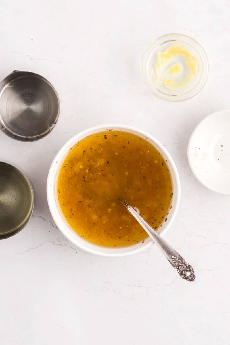 A bowl of vinaigrette with a spoon inside, surrounded by empty measuring cups and a small dish of mustard on a white surface.
