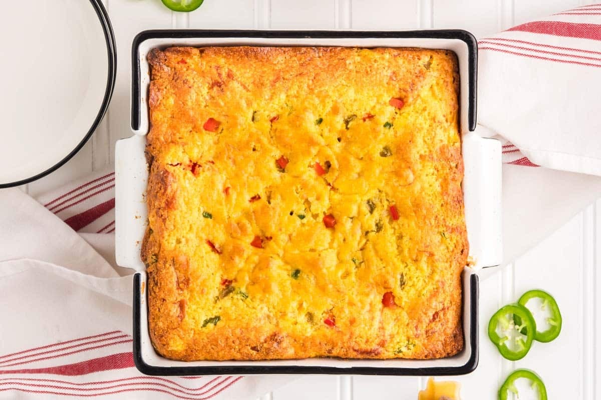 A baked casserole dish of golden Jiffy Mexican cornbread with visible pieces of red and green peppers, set on a white surface with red-striped towels and sliced jalapeños nearby.