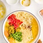 A bowl contains eggs, shredded cheese, chopped red and green peppers, cornmeal, and seasonings, with various small bowls and ingredients arranged nearby on a white surface.