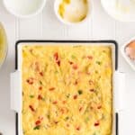 A square baking dish filled with unbaked cornbread batter mixed with chopped vegetables, surrounded by small bowls and baking ingredients on a white surface.