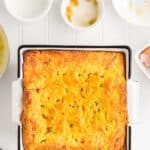 A baked Jiffy Mexican cornbread in a square dish, surrounded by small bowls containing milk, ingredients, and eggs on a white surface.