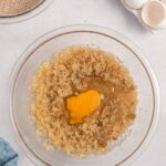 A glass bowl with creamed sugar and butter topped with an egg, next to a bowl of cocoa powder and a carton of eggs on a white surface.