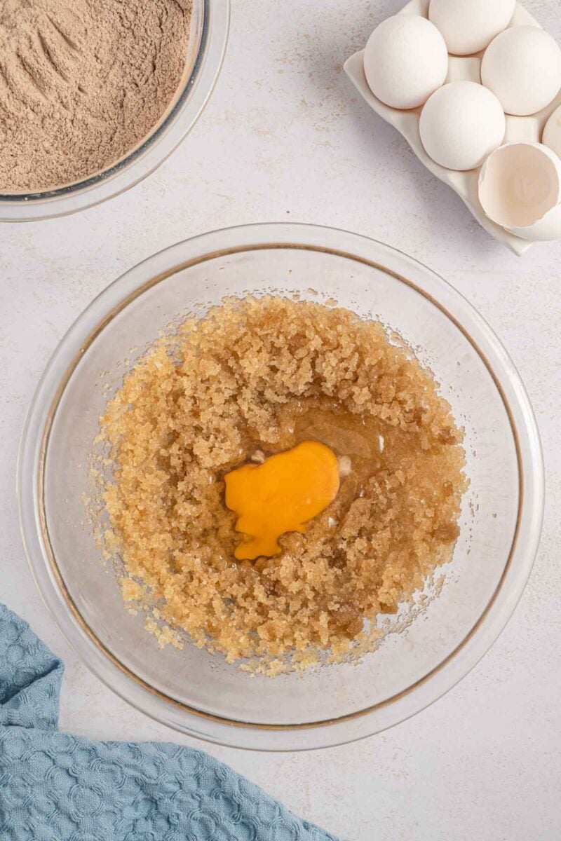 A glass bowl with creamed sugar and butter topped with an egg, next to a bowl of cocoa powder and a carton of eggs on a white surface.