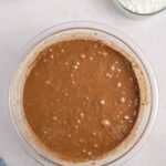 A glass bowl filled with chocolate batter sits on a counter, with a bowl of mini marshmallows and a bowl of powdered sugar nearby.