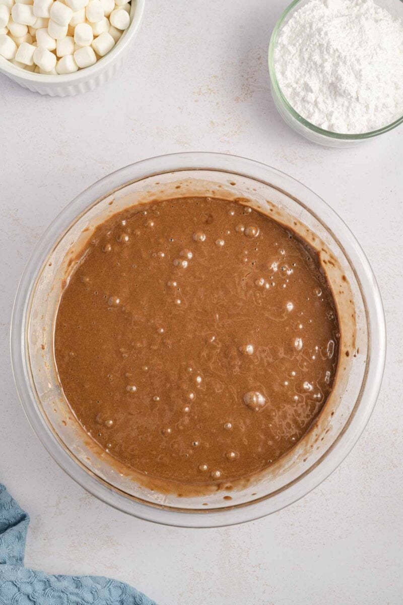 A glass bowl filled with chocolate batter sits on a counter, with a bowl of mini marshmallows and a bowl of powdered sugar nearby.