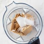 Overhead view of a blender containing broken graham crackers and granulated sugar on a white countertop.