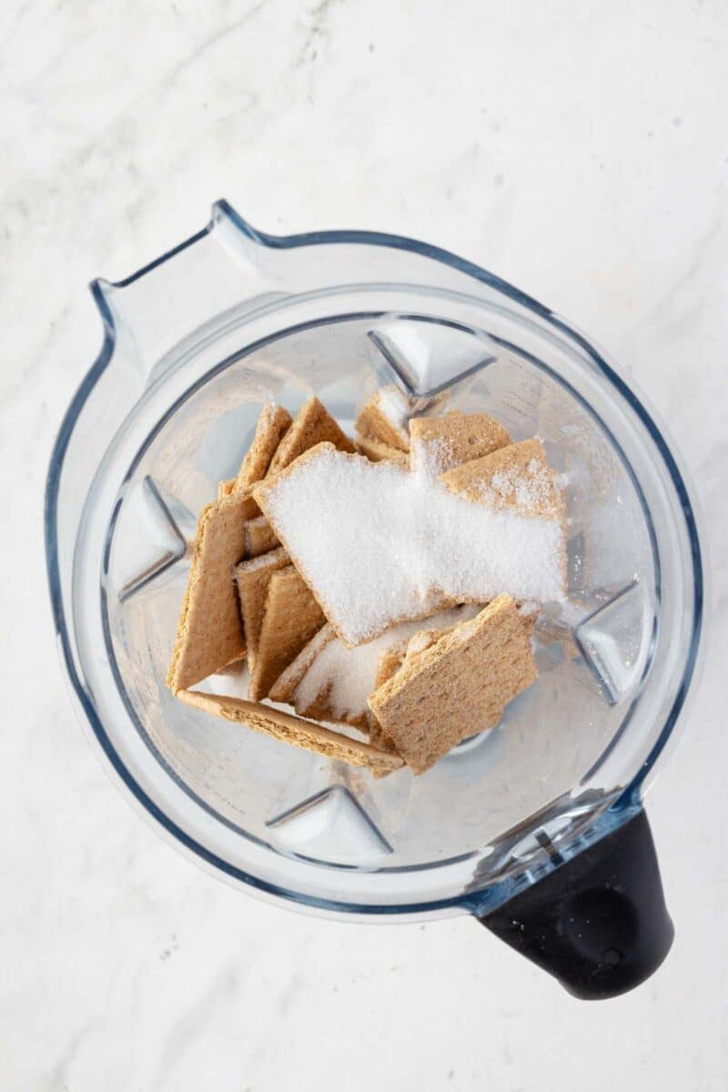 Overhead view of a blender containing broken graham crackers and granulated sugar on a white countertop.