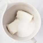 A white mixing bowl containing two blocks of cream cheese and a mound of granulated sugar on a marble surface.