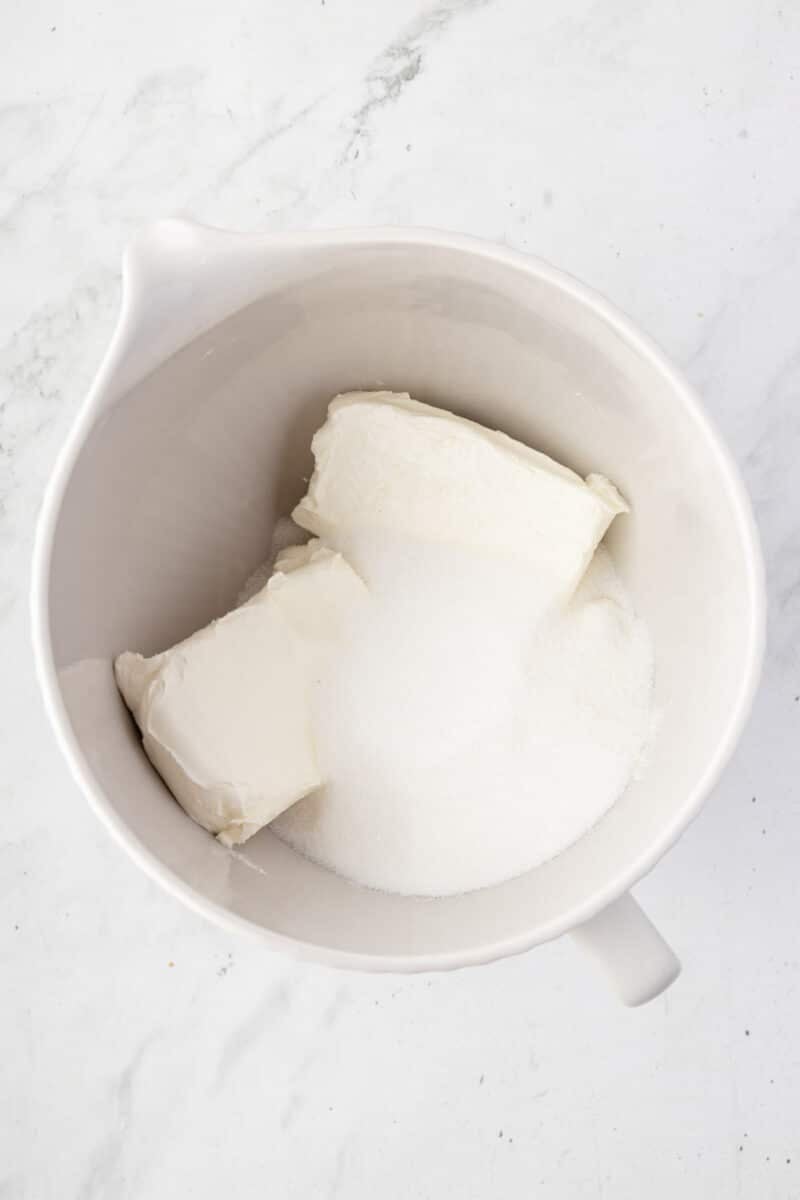 A white mixing bowl containing two blocks of cream cheese and a mound of granulated sugar on a marble surface.
