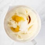 A mixing bowl containing creamed butter and sugar with two raw eggs and a small amount of vanilla extract on top, ready to be mixed.