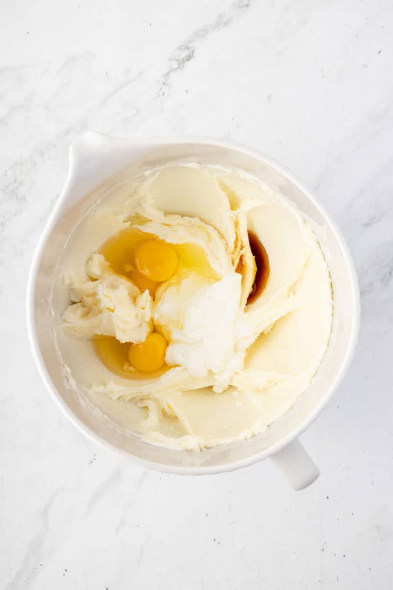 A mixing bowl containing creamed butter and sugar with two raw eggs and a small amount of vanilla extract on top, ready to be mixed.