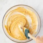 A hand uses a spatula to mix a creamy, orange and white batter in a clear glass bowl on a white surface.