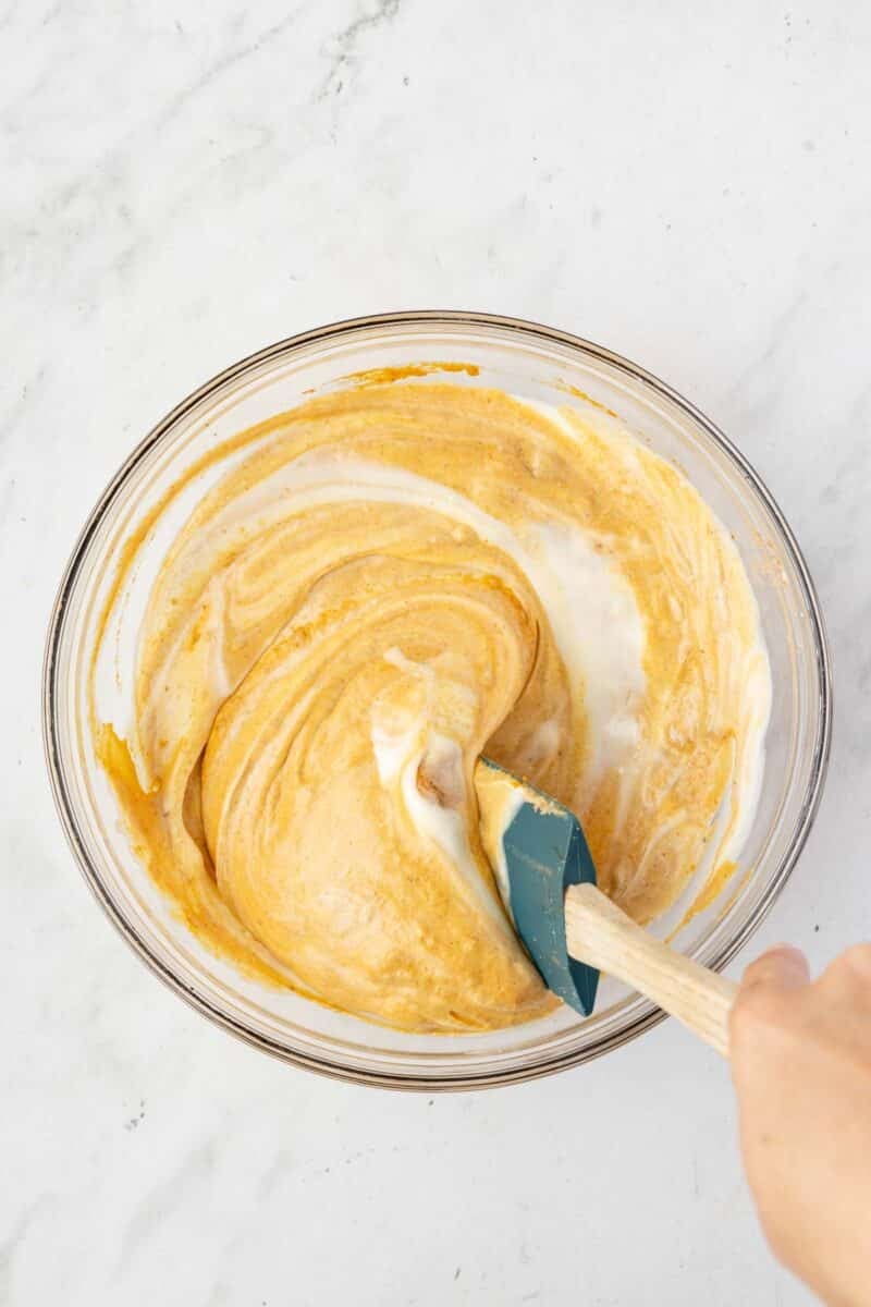 A hand uses a spatula to mix a creamy, orange and white batter in a clear glass bowl on a white surface.