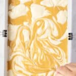 A hand uses a utensil to swirl white and orange batters together in a rectangular baking pan lined with parchment paper.