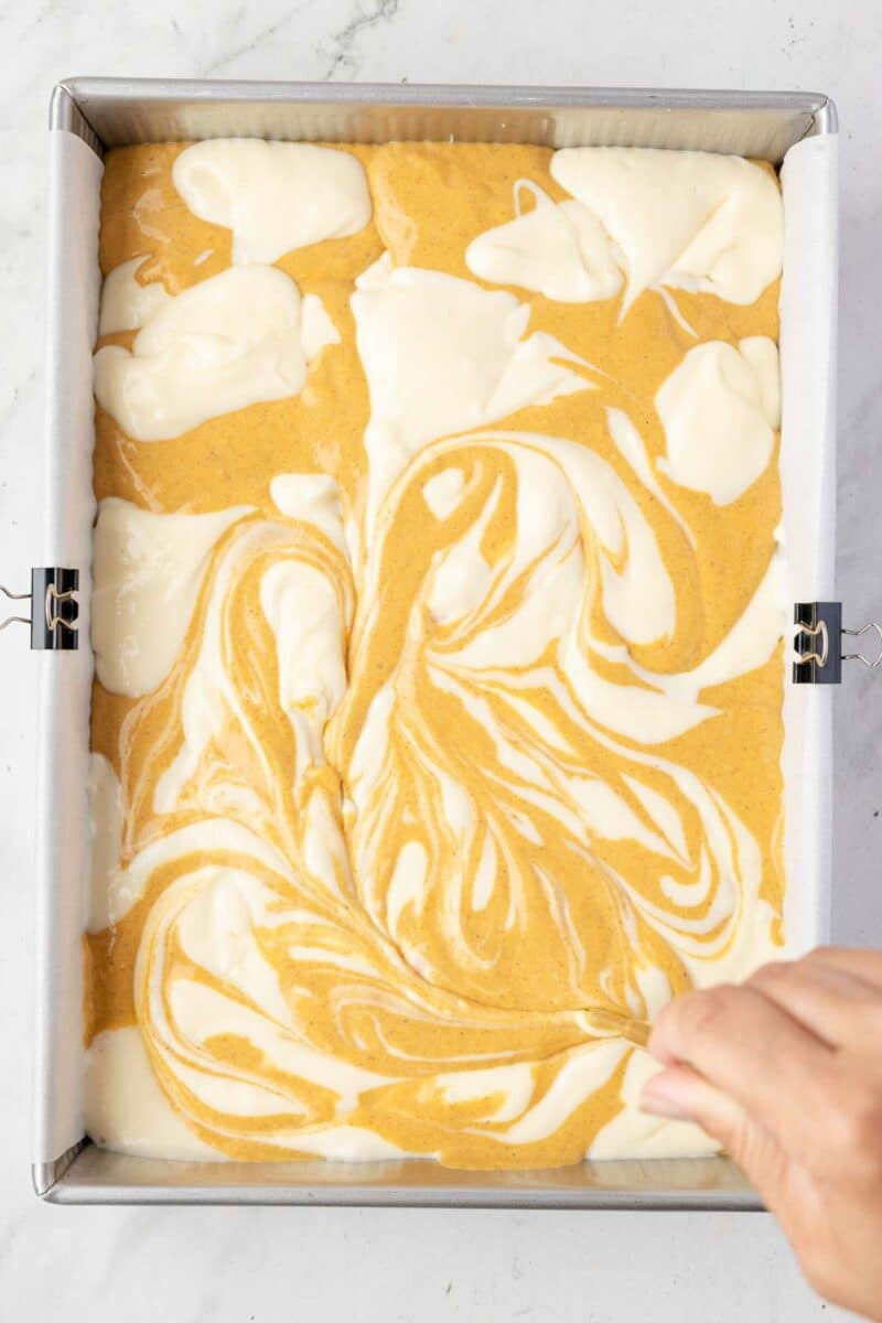 A hand uses a utensil to swirl white and orange batters together in a rectangular baking pan lined with parchment paper.