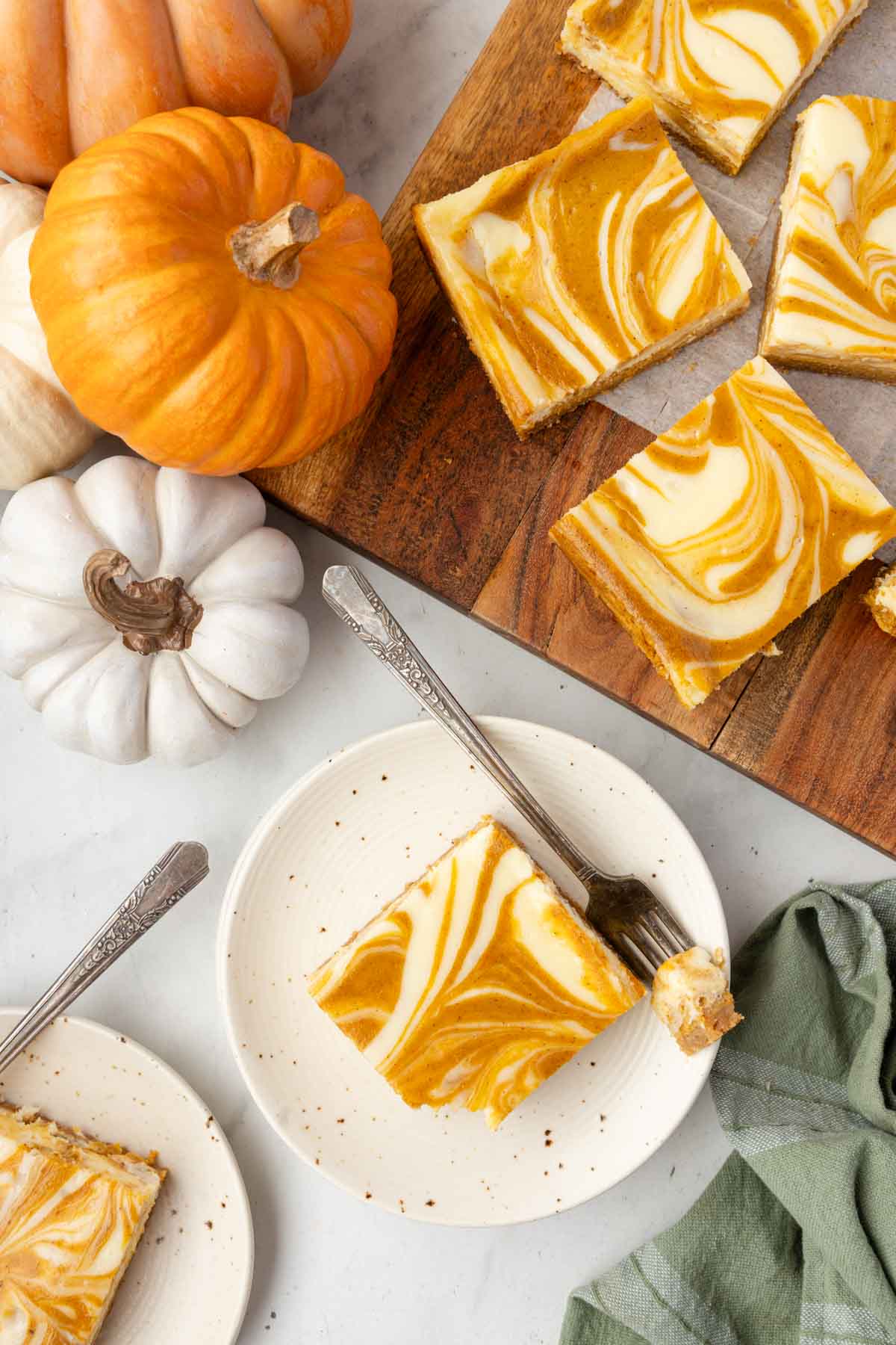 Slices of pumpkin cheesecake bars with swirled tops on plates and a cutting board, surrounded by small pumpkins on a white surface.