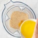 A person pours melted butter from a clear bowl into a blender containing ground graham cracker crumbs on a white surface.