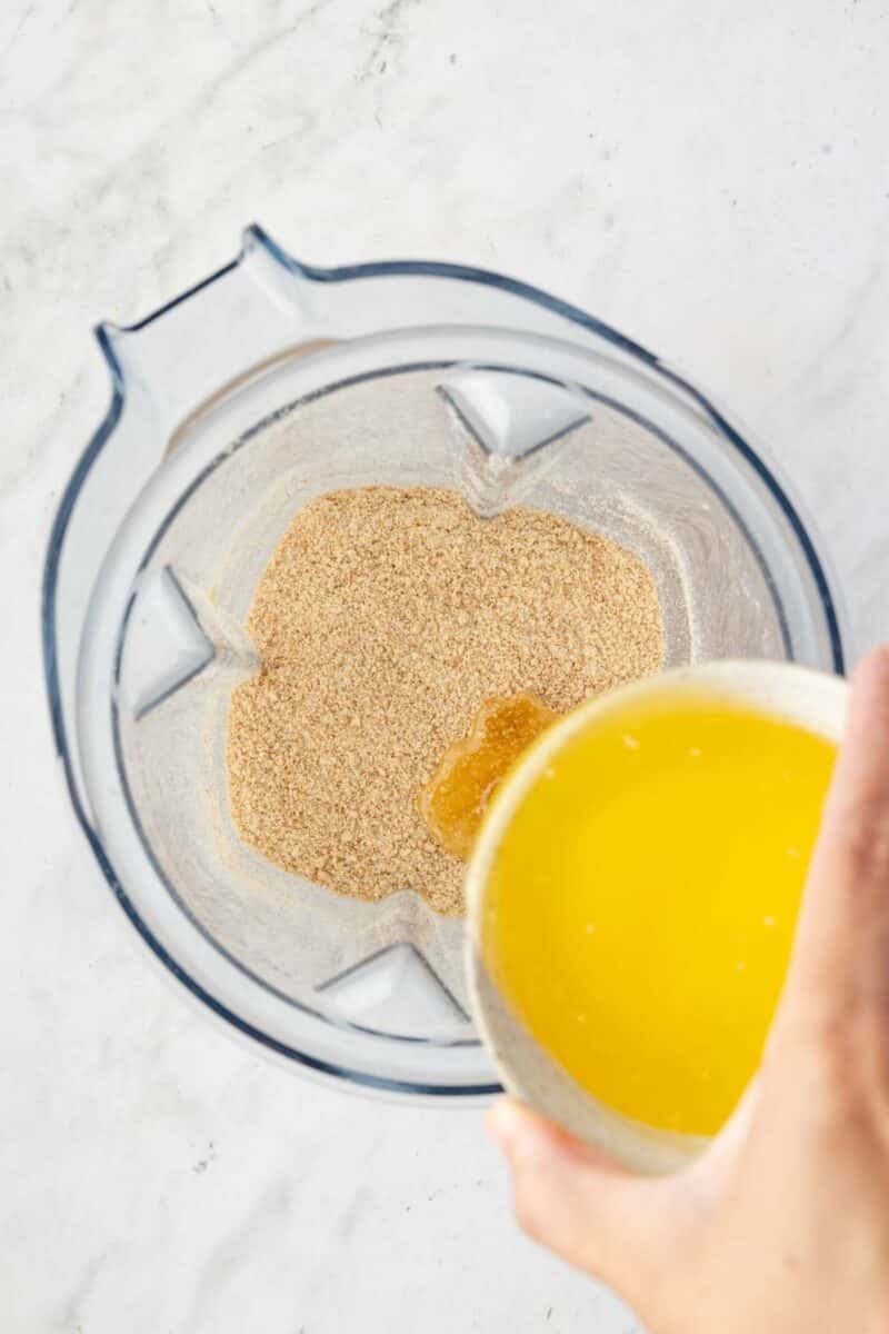 A person pours melted butter from a clear bowl into a blender containing ground graham cracker crumbs on a white surface.