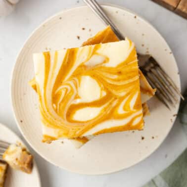 A plate with two pumpkin swirl cheesecake bars stacked, showing a marbled orange and cream pattern, and a fork beside them on a white surface.