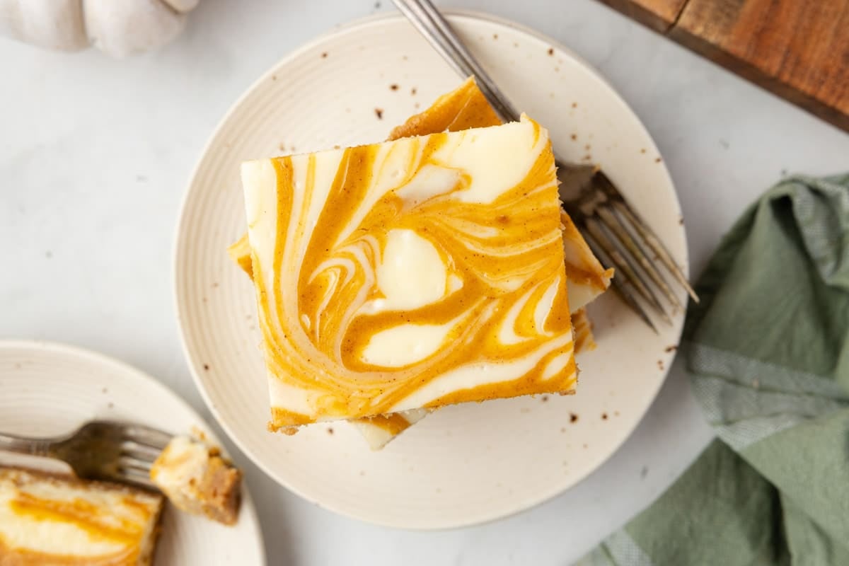 A plate with two pumpkin swirl cheesecake bars stacked, showing a marbled orange and cream pattern, and a fork beside them on a white surface.
