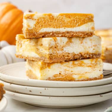 Three pumpkin cheesecake bars are stacked on a plate, with whole pumpkins in the background and a piece of bar on a fork in the foreground.