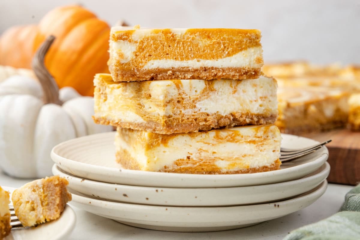 Three pumpkin cheesecake bars are stacked on a plate, with whole pumpkins in the background and a piece of bar on a fork in the foreground.