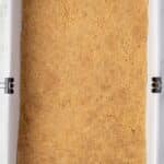 A rectangular baking pan lined with parchment paper, filled with an even layer of pressed graham cracker crust.