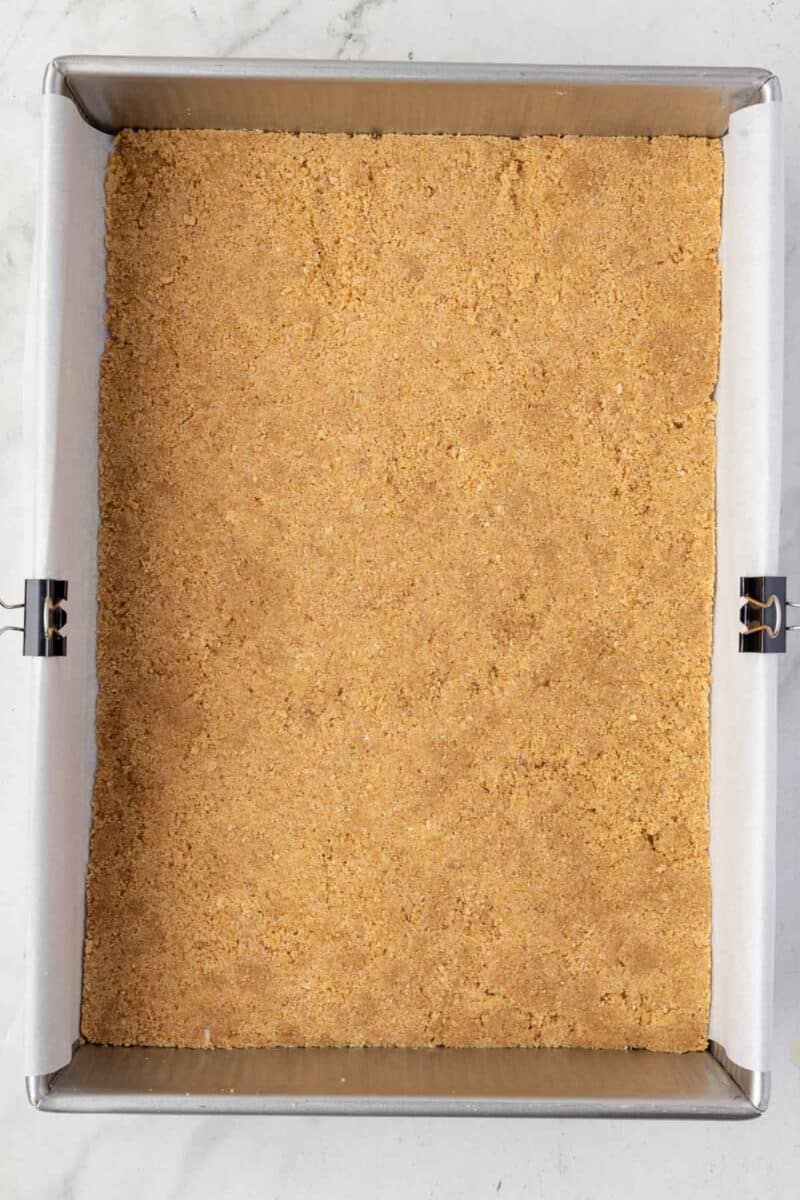 A rectangular baking pan lined with parchment paper, filled with an even layer of pressed graham cracker crust.