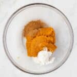 A glass bowl contains pumpkin puree, ground cinnamon, and flour on a white marble surface.